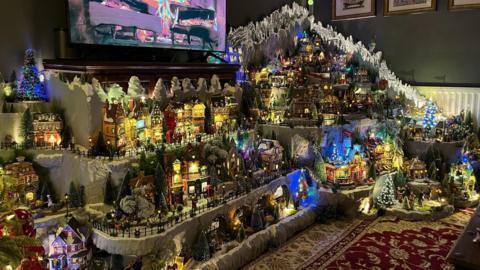 A huge wintery Christmas village in the living room of a house in Bristol. The village is made out of miniature houses with snow, a pub, figurines and lights.