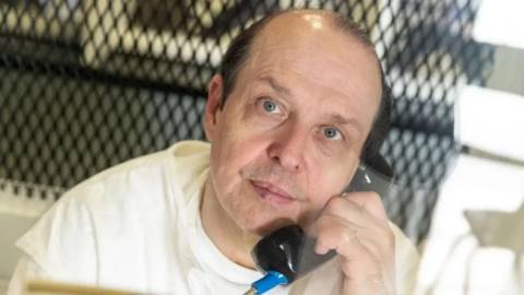 Robert Roberson, in a white prison outfit, looks into the camera with a prison telephone at his ear