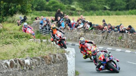 Four racers in colourful leathers lean over while rounding a stone wall on the course, with spectators looking on.