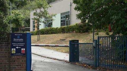 A school gate made of blue metal. A sign next to the gate reads "Sevenoaks Primary School". A school building sits in the background surrounded by trees and hedges.