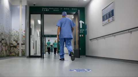 A medical professional wearing blue scrubs walking down a hospital corridor 