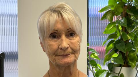 A mature woman with short, light hair is pictured indoors, dressed in a blue and white patterned blouse and wearing a necklace. Behind her, vertical blinds cover a window that lets in natural light, and a leafy green plant sits to the right, adding a touch of nature to the setting.