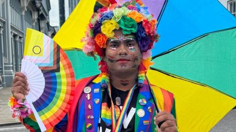 A man stands in the street in a rainbow suit, holding a rainbow fan in his right hand and a rainbow umbrella in his left that is open behind his back. He also has a head dress made of rainbow coloured flowers and festival makeup