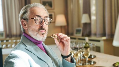 A stern looking man with grey hair holds a macaron; he wears a lilac suit and black rimmed glasses