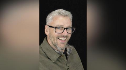 Craig Hurcombe smiles at the camera. He has short grey hair and is wearing black thick-rimmed glasses and a brown shirt. He is pictured against a black background.