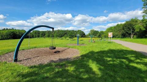 An empty play park on a green with minimal play equipment. The image was taken on a sunny day and trees can been seen in the background.