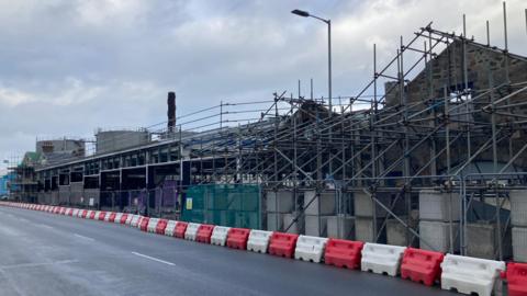 Scaffolding and fencing around a building site in Guernsey. The site appears to be a set of old buildings being renovated. Red and white bollards are on the road next to the fencing.