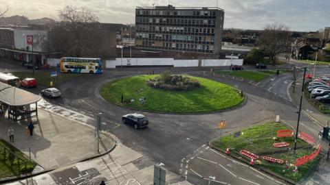 Shot from above of a roundabout covered in grass, with a sculpture in the centre. There are cars going round the roundabout. There is a six-storey building on the far side of the roundabout, and smaller buildings to the left.