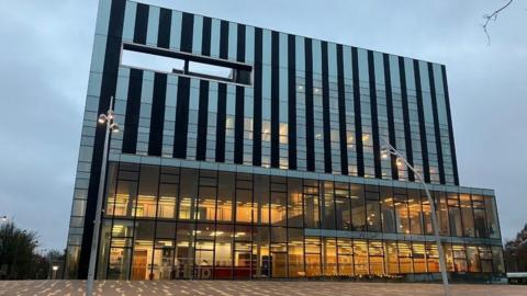 North Northamptonshire Council's headquarters in The Cube at Corby. A large building with glass panels covering its exterior can be seen. Lights are on in the lower half of the building. 