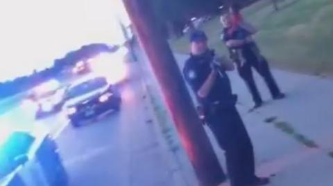 Still from Facebook Live feed shows police car and police officer near it pointing gun at the woman - another police officer is behind him, holding a young girl - July 2016