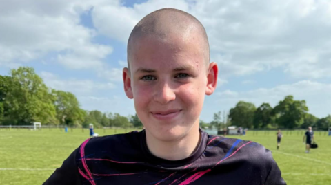 Brodan has a shaved head and wearing a dark football top and is standing in a field or park with people playing behind him. He is smiling at the camera. The sun is shining.