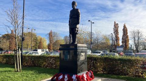 A bronze figure of a male soldier wearing a turban stood on a plinth, which has poppy wreaths placed at its base. There is a green hedge and a car park behind it.