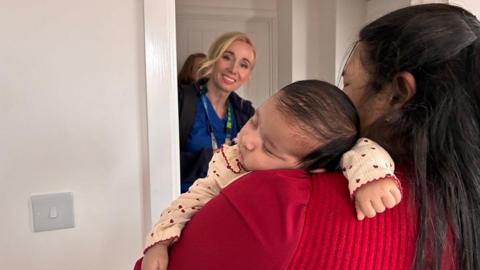 Emma, a health visitor, in her blue uniform is smiling at a woman in a red top with long black hair. A baby is asleep on the woman's shoulder with her arms outstretched. 
