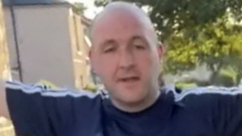 A bald man stands with his arms raised in a residential street. His mouth is open but his face is expressionless. He is wearing a navy blue Adidas t-shirt with white stripes on the sleeve.