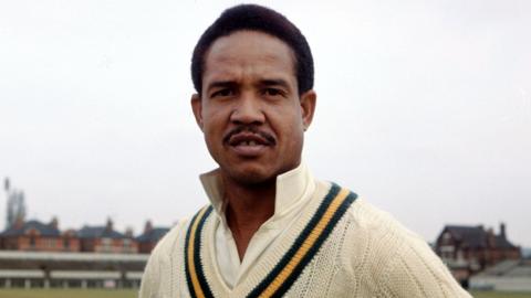 A colour head-and-shoulders shot of a man in his 30s in cricketing attire, a white cricket top with a black-and-yellow stripe. He is standing facing the camera in an empty cricket ground on an overcast say. He has short black hair and a thin black moustache.