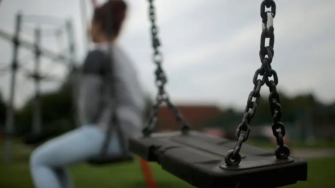 A teenager sits on a swing in a park with her back to the camera. She is wearing jeans and a grey jumper. An empty swing is in the foreground of the image.