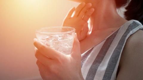 A woman in a stripy top holding a glass of ice water and touching her neck.