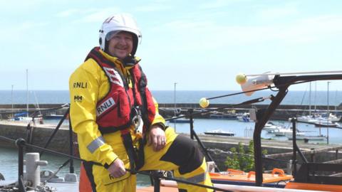 Brendan Rooney is smiling at the camera, wearing a yellow lifeboat crew coat and trousers and a red lifejacket. He has a white helmet on and is leaning on a black fence at a harbour with boats and the sea in the background.