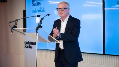 A man wearing black glasses and a dark suit stands in front of a lectern that has Goldman Sachs written on it. He is clasping his hands together. Behind him is a large TV screen