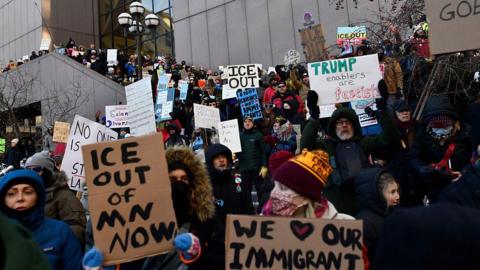 A crowd of protesters waving anti-ICE signs and wearing winter clothes stand on the stairs of a large concrete building in Minnesota