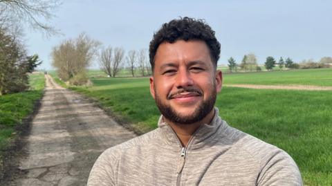 Ben Brooke has short, neat dark hair, a bit curly on top, he has a matching beard and moustache and is wearing a grey cotton sports top zipped up to his neck. Behind him are grass fields with a rough track wide enough for one vehicle running through it.