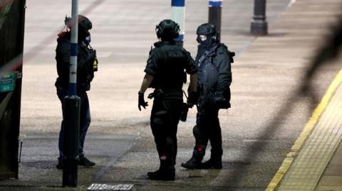 Three specially trained officers dressed in dark clothing, helmets and gloves stand on a train station platform