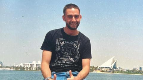 A smiling person sits casually on a railing with both thumbs up, wearing a black graphic T-shirt and blue jeans.