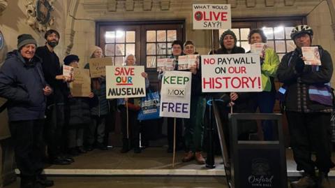 Ten protesters gather at the top of the entrance steps of the town hall. Placards include 'save our museum' and 'no paywall on our history'.