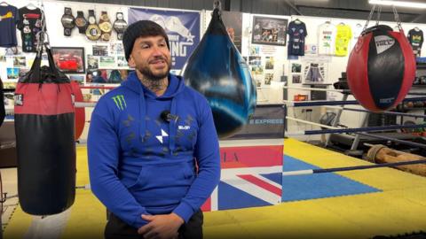 Darren in a gym. He is being interviewed and wearing a mic. There is a ring behind him. Punching bags are hanging up around him. 