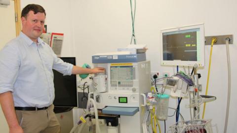 A man with short dark hair and wearing a light blue shirt is standing next to - and resting his left hand on part of - a large medical monitoring item, with wires, leads and pumps and a screen to the right. 