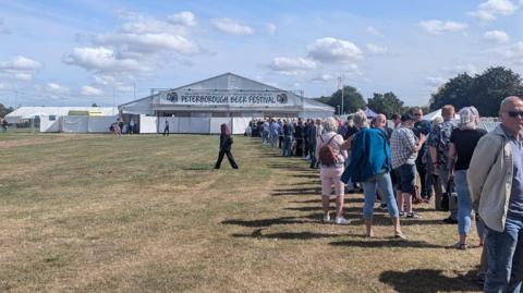 On a sunny day, a long queue of people stretches towards a distant marquee bearing a sign that reads "Peterborough Beer Festival"