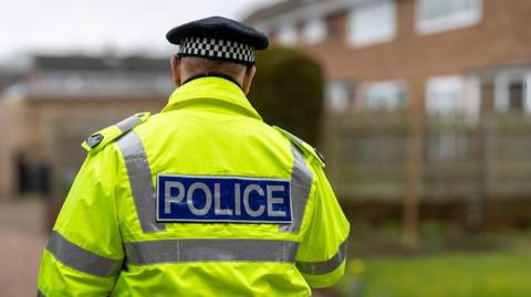 The back of a police officer in a hi-vis coat and black hat standing in front of some houses.