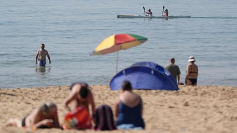 People on beach during May heatwave. One man wades in the sea whist two people paddle by in a kayak