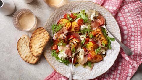 A plate of grilled peaches, mozzarella and tomatoes with silver serving spoons and a red cloth