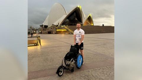 Billy Whiston is standing with his wheelchair in front of the Sydney Opera House, at the start of his journey.