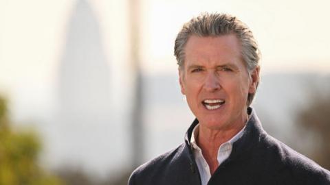 Gov Gavin Newsom stands before a brightly lit city landscape and some trees