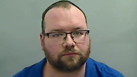 Andrew Barber, who wears black glasses and has a short beard, looks up at the camera. He wears a blue shirt and has no expression. His short dark hair has receded to the top of his head.