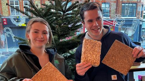 Caelo and Hugo stand indoors in front of a Christmas tree, each holding rectangular boards made from compressed Christmas tree material. Both are smiling at the camera.