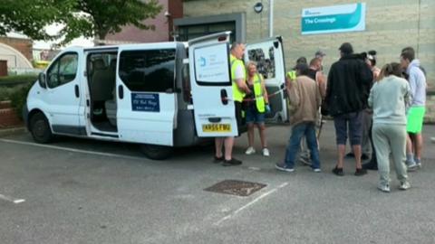 ˿less people queue up to receive food and drink from the back of a white Help the ˿lezz van