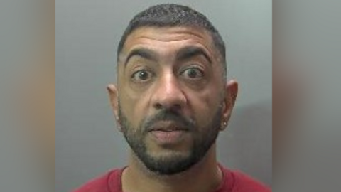 Custody image of Ali Amjad against a grey background. He has an earing in his right ear and dark short hair and a dark beard. He is wearing a red top. His eyebrows are raised so he appears shocked.
