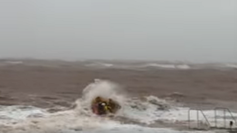 Image shows lifeboat crew heading out on a windy day to rescue two wingsurfers. The volunteer crew, dressed in yellow, are on a small orange boat. They are heading out into stormy seas. Large waves are crashing over the boat carrying the crew.