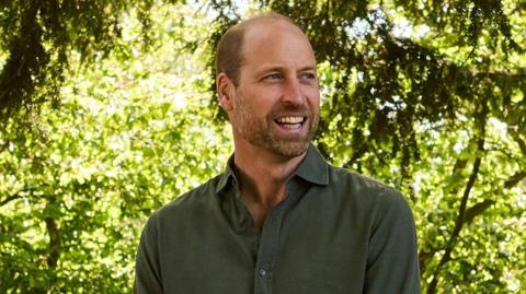 Prince William is wearing a dark green shirt. He is looking away from the camera and smiling. The background is out of focus but has greenery and trees in the background. 