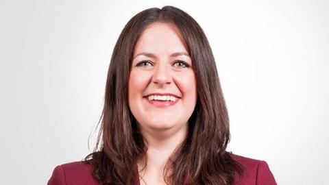 A headshot of Laura. She has long brown hair, is wearing a maroon jacket, and is smiling into the camera.
