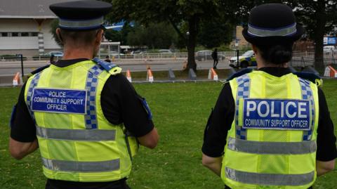 A man and a woman standing in a park, wearing high-vis jackets, with blue plastic logos on the back which says 'police, community support officer'.