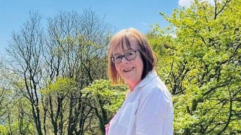 Annie Kissack, who has short, bobbed brown hair, wears glasses. She is smiling for the camera as she poses for a photograph outdoors, with several trees behind her.