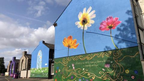 Murals on the end of rows of terraced houses feature grass, blue sky and various flowers in different colours including orange, pink and blue.