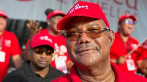 A head and shoulders shot of Patrick Herminie. he is smiling and wearing a red campaign baseball cap. Supporters can be seen behind him - they are out of focus.