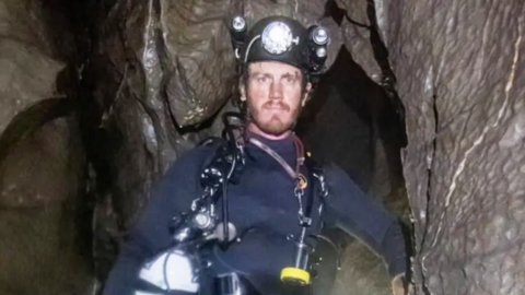 George Linnane inside a cave and looking at the camera. He is wearing a wetsuit and a safety helmet.