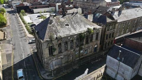 A large two storey grey stone building on the corner of a street is surrounded by fencing on the ground floor/ Trees and plants are growing through holes in the windows and the roof.