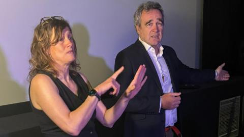 Dr Chris Reid and British Sign Language interpreter, Helen from Hearing Loss Cornwall, are standing in front of a cinema screen. He is wearing a white shirt and black jacket and is gesturing as he speaks. She is wearing a black top and has a pair of glasses on top of her head and she is signing what he is saying.